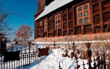 W tle drewniany kości&oacute;ł, przed kościołem trawa pokryta śniegiem obok płotek 