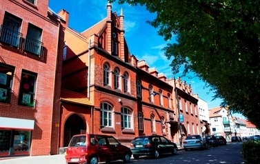 Fragment ulicy przy kt&oacute;rej z budynkami z czerwonej cegły, na ulicy zaparkowane samochody