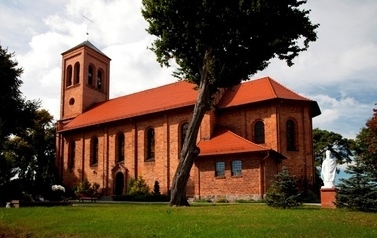 Na pierwszym planie budynek kościoła z czerwonej cegły, wok&oacute;ł budynku drzewa