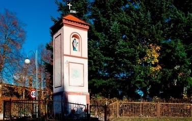 Murowana kapliczka z krzyżem i figurką Jezusa ogrodzona metalowym płotem, za nią widoczne rozłożyste drzewo, z lewej strony zdjęcia latarnia świetlna i znak ograniczenia prędkości do 40