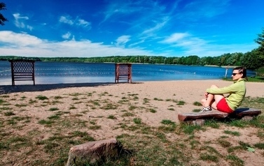Fragment plaży i jeziora, za jeziorem w oddali drzewa, po prawej strony zdjęcia na ławeczce kobieta w okularach przeciwsłonecznych, ubrana w zieloną bluzę i czerwone spodenki