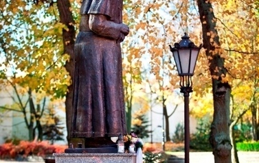 Fotografia przedstawia pomnik na granitowym postumencie stojący w parku, przed pomnikiem leżą wiązanki kwiat&oacute;w, obok stoi niewielka latarnia, liście na drzewach mają barwy pomarańczowo-ż&oacute;łte