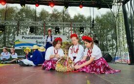 Plener; na scenie dziewczynki w biało czerwonych strojach; na głowach mają wianki; przedstawiają obrzęd dożynkowy.