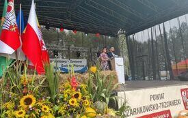 Plener; na scenie stoją prowadzący-kobieta i mężczyzna; z lewej strony powiewają flagi powiatu i Polski, przed sceną snopek siana w otoczeniu słonecznik&oacute;w i dyń.