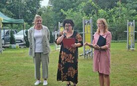 Plener, na pierwszym planie trzy kobiety; każda z nich w ładnym stroju; witają przybyłych na olimpiadę gości i uczestnik&oacute;w.