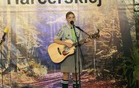 Pomieszczenie; na pierwszym planie młoda dziewczyna ubrana w str&oacute;j harcerski; gra na gitarze; śpiewa do mikrofonu.