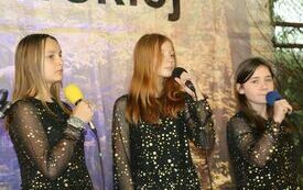 Pomieszczenie; na pierwszym planie trzy młode dziewczyny ubrane w r&oacute;wne, czarne sukienki. Wszystkie trzymają w rękach mikrofony. Przygotowują się do śpiewania.