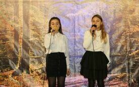 Pomieszczenie; na pierwszym planie dwie młode dziewczyny, ubrane w stroje galowe, śpiewają piosenkę trzymając w rękach mikrofony.