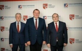 Pomieszczenie; na pierwszym planie trzech mężczyzn; wszyscy odświętnie ubrani; w tle herb powiatu czarnkowsko-trzcianeckiego oraz logo Forum Gospodarczego.