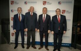 Pomieszczenie; na pierwszym planie czterech mężczyzn; wszyscy odświętnie ubrani; w tle herb powiatu czarnkowsko-trzcianeckiego oraz logo Forum Gospodarczego.