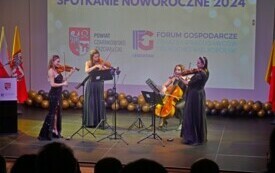 Pomieszczenie; na scenie cztery kobiety; trzy z nich grają na skrzypcach, jedna na wiolonczeli; w tle herb powiatu i logo Forum Gospodarczego. Na posadzce leżą czarno-złote balony.