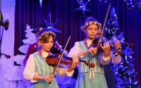 Pomieszczenie; na scenie dwie dziewczynki w jasno zielonych sukienkach, na głowach wianki z małych, kolorowych kwiatk&oacute;w; obie grają na skrzypcach.