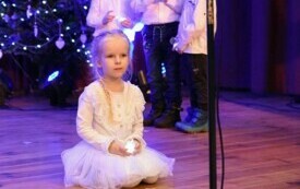 Pomieszczenie; na pierwszym planie klęcząca na scenie dziewczynka, ubrana cała na biało, włosy splecione w warkocz, w rękach trzyma świeczkę.