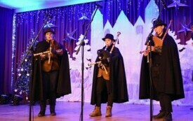 Pomieszczenie; na scenie trzech młodych chłopak&oacute;w, każdy z nich gra na dudach, ubrani w długie, czarne peleryny i kapelusze na głowie.