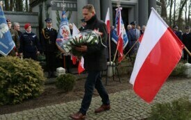 Plener; na pierwszym planie mężczyzna niosący wiązankę z białych kwiat&oacute;w. Za nim powiewające flagi biało-czerwone oraz inni uczestnicy wydarzenia.