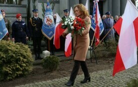 Plener; na pierwszym planie kobieta w beżowym płaszczu niosąca wiązankę złożoną z biało-czerwonych kwiat&oacute;w, przewiązaną białą i czerwoną wstążką; w tle uczniowie ze sztandarami; na wietrze powiewają flagi Polski.