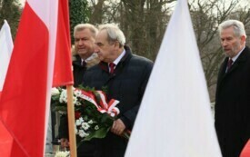 Plener; na pierwszym planie trzech mężczyzn z wiązanką; pomiędzy nimi powiewa flaga Polski. 