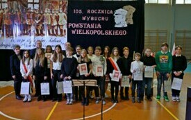 Pomieszczenie; grupa uczni&oacute;w z dyplomami i nagrodami; za nimi baner z napisem 105. rocznica wybuchu Powstania Wielkopolskiego.