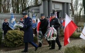 Plener; na pierwszym planie trzech mężczyzn ubranych w mundury; wszyscy trzymają w rękach biało-czerwone wiązanki; w tle flagi Polski oraz inni uczestnicy spotkania.