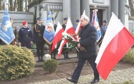 Plener; na pierwszym planie mężczyzna z biało-czerwoną wiązanką kwiat&oacute;w. W tle flagi Polski oraz osoby ubrane w mundury.