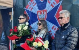 Plener; na pierwszym planie dwie młode kobiety w rękach trzymające biało czerwone bukiety oraz starszy mężczyzna. Za nimi logo WOT. 