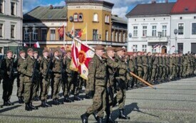 Plener; na pierwszym planie trzech mężczyzn, jeden z nich trzyma sztandar; za nimi pozostali żołnierze stojący na baczność.