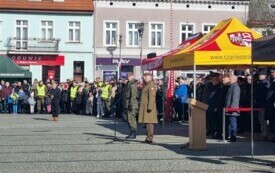 Plener; na pierwszym planie przemawiający pułkownik; za nim władze samorządowe i przedstawiciele służb mundurowych. W tle obywatele przybyli na uroczystość.