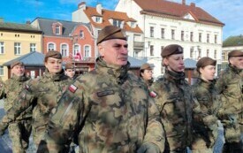 Plener; na pierwszym planie grupa maszerujących żołnierzy; w tle budynki mieszkalne oraz budynek Urzędu Miasta.