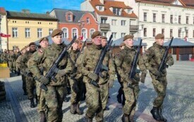 Plener; na pierwszym planie grupa maszerujących żołnierzy; w tle budynki mieszkalne oraz budynek Urzędu Miasta.