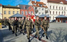 Plener; na pierwszym planie trzech mundurowych maszerujących ze sztandarem; za nimi spora grupa żołnierzy z bagnetami. W tle powiewają biało-czerwone flagi.