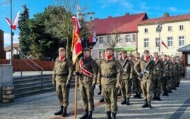 Plener; na pierwszym planie trzech mundurowych stojących ze sztandarem; za nimi spora grupa żołnierzy z bagnetami. W tle powiewają biało-czerwone flagi.