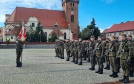 Plener; naprzeciwko sztandaru stoją żołnierze ubrani w mundury; wszyscy stoją na baczność; w tle powiewają biało-czerwone flagi; w tle kości&oacute;ł p.w. Marii Magdaleny w Czarnkowie.