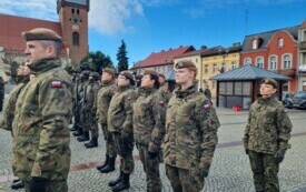 Plener; na pierwszym planie panie w mundurach; wszystkie stoją na baczność. W tle budynki mieszkalne oraz kości&oacute;ł p.w. Marii Magdaleny w Czarnkowie.