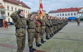 Plener; W szeregu stoją żołnierze w mundurach. Jeden z nich trzyma sztandar, pozostali w rękach mają karabiny, inni salutują. W tel budynki mieszkalne i budynek Urzędu Miasta.
