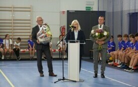Pomieszczenie; przy m&oacute;wnicy kobieta z dwoma mężczyznami. Jeden z nich w rękach trzyma bukiet kwiat&oacute;w, drugi kosz, w kt&oacute;rym znajdują się sportowe piłki. Za nimi na sportowych ławeczkach siedzą dzieci ubrane w stroje do ćwiczeń.