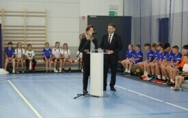 Pomieszczenie; przy m&oacute;wnicy kobieta z mężczyzną; oboje odświętnie ubrani. Za nimi na sportowych ławeczkach siedzą dzieci ubrane w stroje do ćwiczeń.