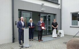 Plener; przed wejjściem do budynku stoją dwie kobiety i dw&oacute;ch mężczyzn, jeden z nich przemawia z m&oacute;wnicy, drugi trzyma otwarty grawerton. Wszyscy elegancko ubrani.