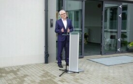 Plener; przed wejściem do budynku ustawiona m&oacute;wnica, przy kt&oacute;rej przemawia mężczyzna w granatowym garniturze.