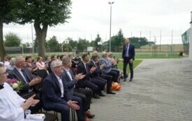 Plener; na pierwszym planie zaproszeni na uroczystość goście; jeden z mężczyzn stoi, obracając się do zebranych. W tle szkolne boisko sportowe.