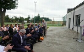 Plener; na pierwszym planie zaproszeni na uroczystość goście; jeden z mężczyzn stoi, obracając się do zebranych. W tle szkolne boisko sportowe.