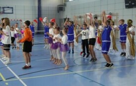 Pomieszczenie; na pierwszym planie dzieci skaczące do g&oacute;ry; wszyscy w r&oacute;żnorakich sportowych strojach. Niekt&oacute;rzy trzymają biało-czerwone pompony w rękach.