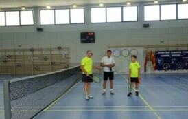 Pomieszczenie; na pierwszym planie dw&oacute;ch mężczyzn w sportowych strojach i chłopiec. Trzymają w rękach rakiety tenisowe. Za nimi baner z olimpijskimi k&oacute;łkami. Nad nimi zawieszona tablica odliczająca punkty.