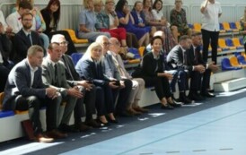 Pomieszczenie; na pierwszym planie odświętnie ubrani zaproszeni na uroczystość goście. Za nimi na trybunach siedzi więcej os&oacute;b. Krzesełka na trybunach są niebieskie i ż&oacute;łte. 