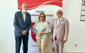 Pomieszczenie, stoją trzy osoby - mężczyzna ubrany w ciemną marynarkę i spodnie, kobieta ubrana w jasnozieloną bluzkę i beżowe spodnie oraz mężczyzna w jasnym garniturze. Fotografowane osoby stoją na tle ścianki promocyjnej z napisem &quot;POWIAT Czarnkowsko-T