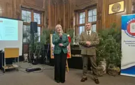 Pomieszczenie. Na pierwszym planie trzech chłopc&oacute;w ubranych w zielone mundury. Na każdym z nich przypięta czerwona wstążka. W rękach trzymają rogi myśliwskie. Za ich plecami ekran wyświetlający prezentację.