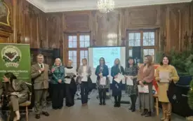 Pomieszczenie, na pierwszym planie cztery dziewczyny śpiewające do mikrofon&oacute;w. Przed nimi statyw z nutami. Za nimi trzech chłopc&oacute;w grających na instrumentach. W tle choinki.