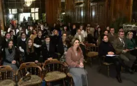 Pomieszczenie, duża sala z drewnianymi ścianami. Na krzesłach siedzą uczestnicy wydarzenia. Część z nich ubrana w mundury leśne. Za ich plecami ściana wyłożona lustrami. Na suficie żyrandol.