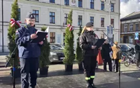 Na placu, stoją przy mikrofonach policjant oraz kobieta ze straży pożarnej. Trzymają w rękach teczki, z kt&oacute;rych czytają przem&oacute;wienie. 