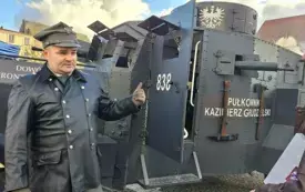 Mężczyzna w mundurze stoi na placu, a za nim znajduje się powojenny czołg.