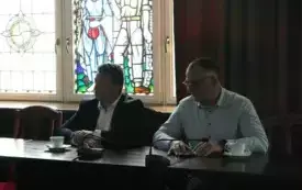 Pomieszczenie; przy stole siedzi dw&oacute;ch mężczyzn. Przed każdym z nich znajdują się mikrofony oraz filiżanki ze spodkiem. W tle widoczna jest witrażowa szyba.